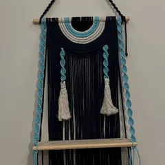 I love creating pieces that are both beautiful and useful. This evil eye themed wall shelf does just that, combining a protective symbol with a practical wooden shelf. The deep navy blue and sky blue cords with twisted hangers make it a unique piece to display small plants or treasures.
