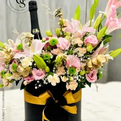 A sophisticated floral hamper featuring a bottle of champagne emerging from a beautiful arrangement of pink lilies, roses, and carnations. It's all presented in a chic black hatbox with a gold ribbon.