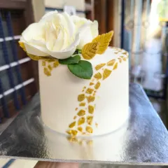 The pristine combination of white and gold. This minimalist cake features a smooth white finish, a textured gold mosaic pattern, and a single, beautiful white sugar rose.