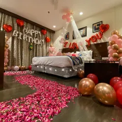 A romantic birthday surprise filling a hotel room with love. The decor includes a rose petal pathway, a canopy with fairy lights over the bed, 'I Love You' and 'Happy Birthday' foil balloons, and balloon arrangements in red and rose gold.