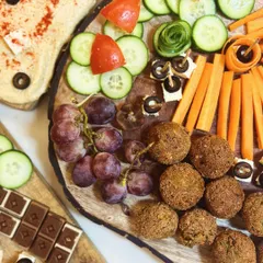 A top-down view of a Mediterranean-inspired grazing board. This platter features homemade falafel, creamy hummus, fresh grapes, and artfully arranged vegetable sticks like carrots and cucumber.