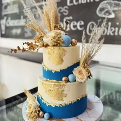 A modern two-tiered cake perfect for a stylish celebration. This design combines a rich royal blue base with an ivory top tier, accented with edible gold leaf, dried pampas grass, and delicate sugar flowers.