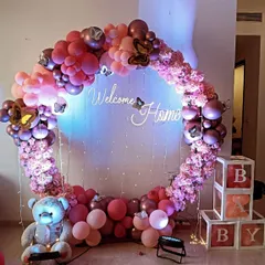 A beautiful circular balloon and floral arch in shades of pink for a welcome home party. This design includes a 'Welcome Home' neon sign, a teddy bear, and 'BABY' blocks, creating a complete and festive photo opportunity.