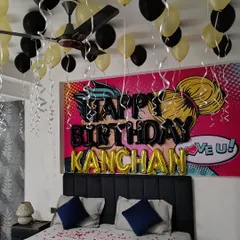 A personalized birthday bedroom surprise with a pop-art backdrop. The decor features black and gold balloons, the name 'KANCHAN' in foil letters, and rose petals scattered on the bed.