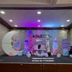 This stage setup for a baby shower features a mix of modern and playful elements. It includes a decorated swing, a 'Baby Shower' neon sign, a crescent moon prop, and large baby bottle cutouts filled with pink and blue balloons.