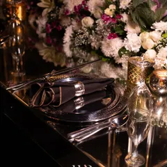 Every place setting is an opportunity for beauty. Here, a silver charger plate and folded black napkin with a silver ring sit on a mirrored table, reflecting a lush arrangement of white and purple flowers.
