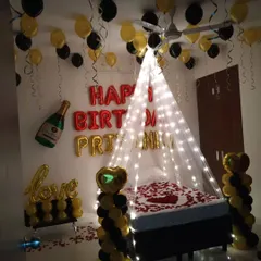 An elegant surprise birthday room decoration featuring a canopy of fairy lights over the bed, rose petals, and a black and gold balloon theme.