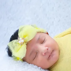 A close-up of a sleeping baby's face, framed by a delicate yellow bow. You can see the soft curve of her lips and the smoothness of her skin.