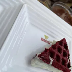 A close-up of our delicious Red Velvet waffle, served on a Vashdev branded plate. The waffle is generously coated with creamy white chocolate, making for an irresistible dessert.