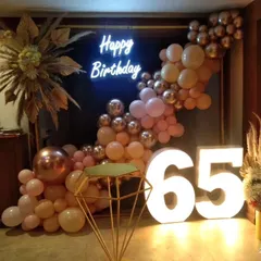 An elegant 65th birthday party setup with a rose gold and pink balloon arch, a "Happy Birthday" neon sign, and large marquee numbers.