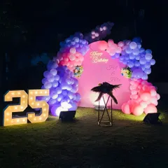 An outdoor 25th birthday celebration at night. The pink backdrop and balloon arch are illuminated by spotlights and the glow of the large marquee numbers.