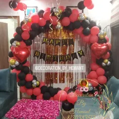 A birthday setup in a living room, featuring a circular backdrop made of red and black balloons. The center table is covered with rose petals, adding a touch of romance and color to the decor.