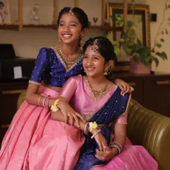 Sisters sharing a happy moment in their coordinated traditional outfits. One wears a pink langa dhavani, the other a matching lehenga.