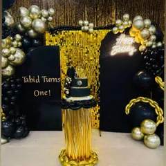 A chic black and gold theme for a first birthday, featuring a shimmer curtain, a custom name board, and a cake placed on a gold pedestal.
