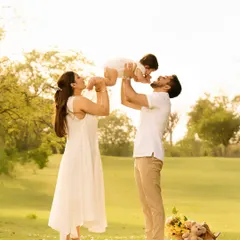 A playful moment captured during a sunny park photoshoot. Both parents lift their baby, their happy expressions and the bright lighting creating a perfect picture of family fun.