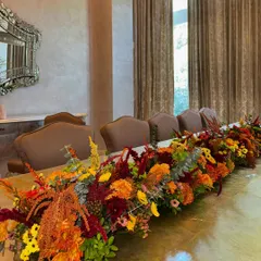 A long, autumnal flower runner designed to dress a dining table for the festive season. The warm tones of orange, red, and yellow create a cozy and inviting atmosphere as the days get shorter and the lights start to twinkle.