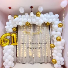 An elegant white and gold balloon arch for a 61st birthday celebration. The fairy light curtain behind the arch adds a warm and sophisticated sparkle to the event.