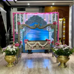 We can transform any space in your home for a ceremony. This is a traditional jhoola setup with a custom-printed backdrop featuring peacock motifs, decorated with pink flowers and placed between two large brass urlis filled with chrysanthemums.