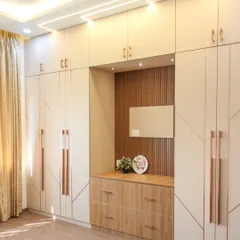 A full view of a large, custom-built wardrobe in a light beige laminate. The design features geometric grooves, long rose gold handles, and an integrated open niche that serves as a compact vanity or display area.