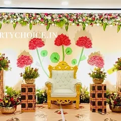 A creative and modern stage for a half saree ceremony. The backdrop features stylized paper flowers and a royal throne, framed by traditional floral pillars.