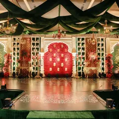 This majestic wedding stage features a symphony of emerald drapes, golden chandeliers, and a central wall of red roses with embedded candles. The design blends traditional Rajasthani architecture with grand floral artistry.