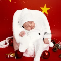 A cozy Christmas-themed newborn photo. The baby is sleeping soundly in a soft white outfit, nestled in a fluffy chair against a festive red background with stars and ornaments.
