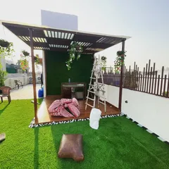 This terrace in Janakpuri features a dedicated relaxation zone under a modern pergola. The space is defined by a wooden deck, a green wall backdrop, and a comfortable lounge setup on the artificial lawn.