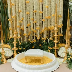 A close-up of a traditional Haldi ceremony space. The setup includes a low white stool placed in a decorative basin, surrounded by golden pillars and a backdrop made of fresh jasmine and marigold strings.