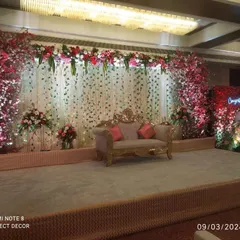 An elegant stage design for a wedding reception. The setup includes a beautiful backdrop of white floral strings, flanked by lush red and white flower arrangements and a classic, ornate sofa for the couple.