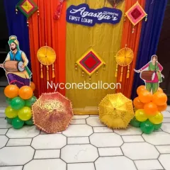 This Lohri backdrop features cutouts of dhol players, adding a traditional Punjabi touch to the celebration. The vibrant orange and yellow drapes, along with decorative umbrellas, create a lively and authentic atmosphere for a first Lohri party.