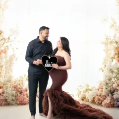 A joyful gender reveal photo for an expecting couple. They are surrounded by a beautiful floral setup as they announce "It's a Girl" with a heart-shaped sign.