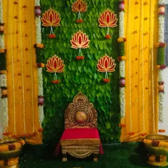 A stunning backdrop for a half saree function, designed with a wall of fresh betel leaves. The setup is framed by yellow drapes and marigold garlands, with a traditional ornate chair placed in the center for the ceremony.