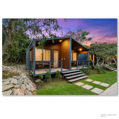A pair of modern prefab cottages at dusk, nestled in a lush mango grove. The warm interior light and the dramatic sunset sky create a magical and inviting scene, showcasing the appeal of modular living.