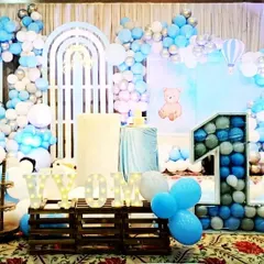 This first birthday stage for Vivom features a charming blue, white, and silver color scheme with a teddy bear theme. The setup includes a large balloon-filled marquee number '1', custom backdrops, and picket fence details for a sweet and classic look.