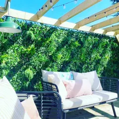 This artificial green wall serves as a beautiful backdrop for an outdoor patio seating area. It is installed under a pergola and adds a natural feel to the space without blocking sunlight.