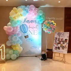 This 'Wonderful One' hot air balloon theme uses a soft pastel color palette. The decor includes a custom arched backdrop, a 3D hot air balloon prop made of balloons and a basket, a neon '1' sign, and a personalized photo collage board for the baby's first year.