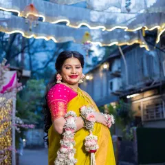 The mother-to-be smiles radiantly, standing under festive outdoor decorations for her baby shower. This shot captures her joy and the celebratory atmosphere of the event, highlighting her traditional attire and floral ornaments.