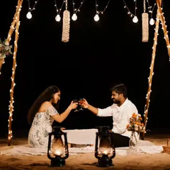 A couple toasts under a canopy of fairy lights on the beach at night. This image showcases a beautifully styled, romantic picnic setup perfect for a pre-wedding shoot.