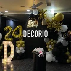A stylish 24th birthday decoration with a black, gold, and white color scheme. The setup includes a large 'D' marquee letter, '24' foil balloons, and a balloon arch against a black backdrop with a neon sign.