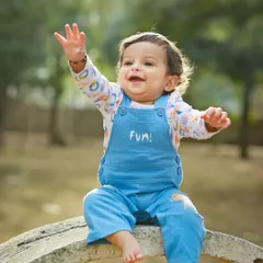 An adorable toddler in blue overalls waves happily during an outdoor photoshoot. The natural setting and the child's joyful expression make for a perfect candid portrait.