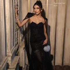 A full view of the look, showing how the sleek top knot bun complements the dramatic black gown and sophisticated makeup.
