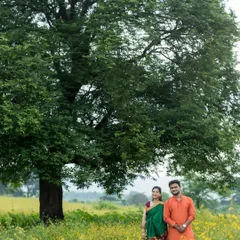A wide shot of an expecting couple in a field of yellow flowers, with a large, majestic tree behind them. Their traditional Indian outfits, a green saree and orange kurta, add a vibrant touch to this classic outdoor maternity portrait.