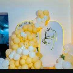 A modern and artistic "He or She" gender reveal or baby shower theme. The setup features a unique arched backdrop with a line-art graphic and a cascade of soft yellow and white balloons.