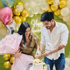 A couple cuts their cake in front of a gold and white balloon backdrop with a 'Happy Anniversary' neon sign. This setup is perfect for photos and celebrating together.
