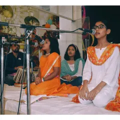 Students performing a group song in traditional attire, accompanied by a tabla player. Group singing helps develop skills in harmony and listening.