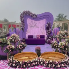 This carnival-themed Mehendi stage features a whimsical purple backdrop with hot air balloon props. The setup includes a deep purple sofa and is surrounded by vibrant floral arrangements, creating a playful and festive atmosphere.