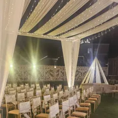 An elegant outdoor ceremony setup with a canopy of white drapes and fairy lights. The aisle is lined with chairs for guests, all facing a simple yet beautiful stage, creating a classic and romantic setting.