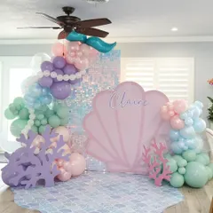 An "Under the Sea" mermaid party with a large pink seashell backdrop and a shimmering sequin panel. The scene is completed with coral props and a pastel balloon garland.