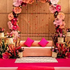 A beautiful indoor Mehendi or Haldi stage decorated with a large arch of paper flowers in shades of pink and peach. The simple sofa and bright pink carpet create a cheerful and elegant space for the ceremony.