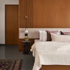 This bedroom at the Amaiti Farmhouse combines a rich wood-panelled headboard wall with soft, neutral bedding. The design is intentionally simple, using a traditional rug and a small, sculptural side table to add layers of personality without clutter.